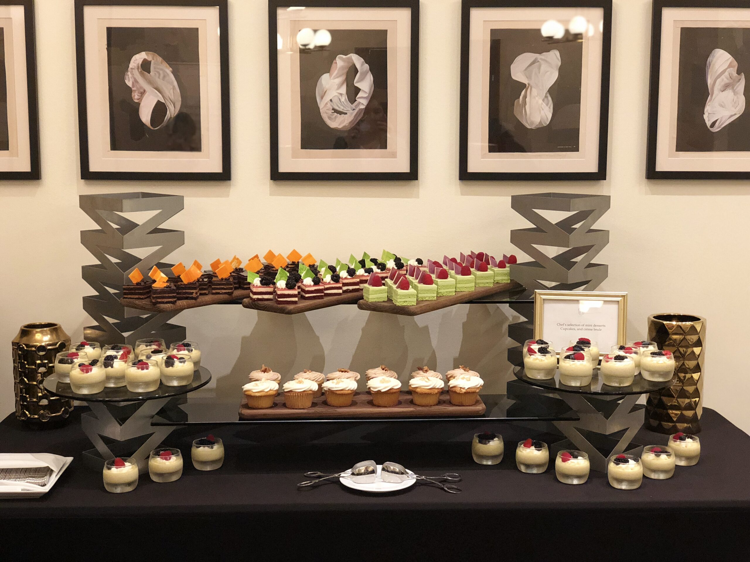A dessert table with assorted cakes, cupcakes, and parfaits, arranged on black stands, with framed abstract art hanging on the wall behind.