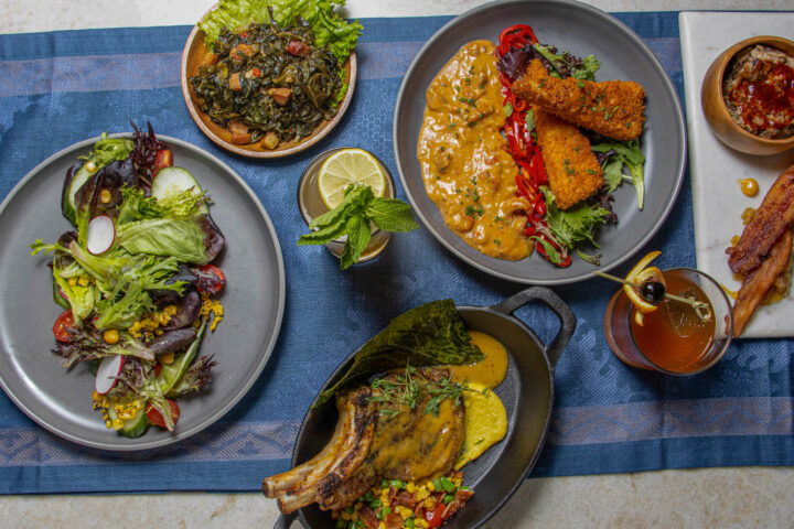 A table set with plates of salad, fried chicken, sauced pork chop, cooked greens, bacon, a drink, and a lemon garnish on a blue table runner.