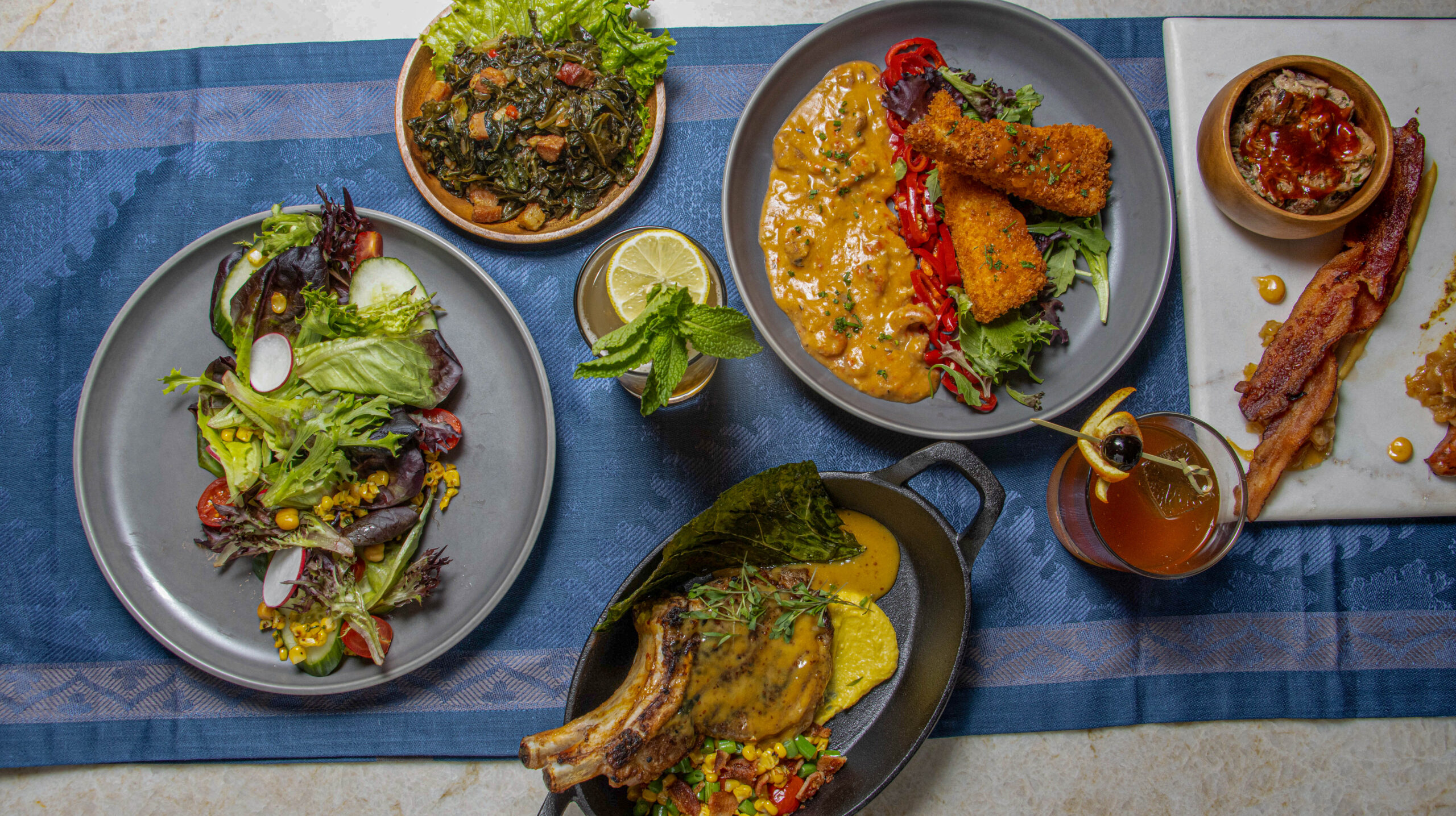 A table set with plates of salad, fried chicken, sauced pork chop, cooked greens, bacon, a drink, and a lemon garnish on a blue table runner.