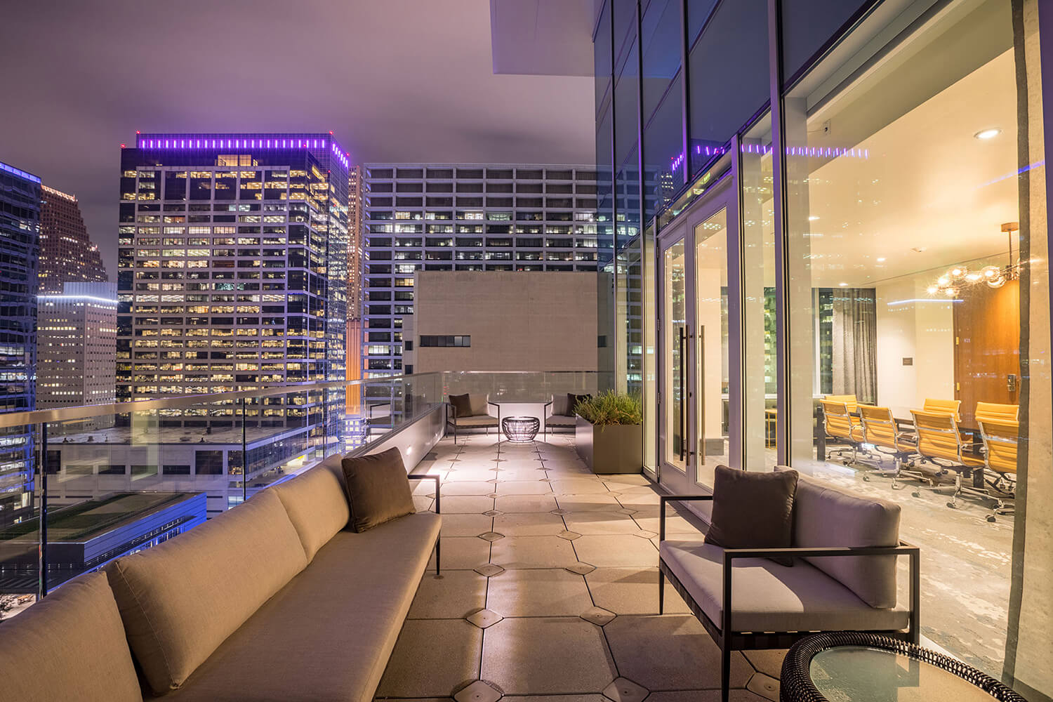 Modern rooftop patio with sofas overlooks city skyscrapers at night; glass doors open to a brightly lit conference room inside.