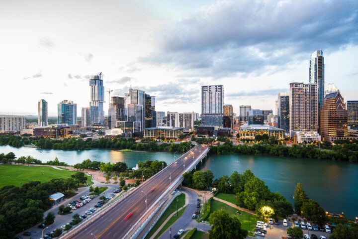 A city skyline with tall buildings, a river, and a bridge surrounded by trees under a cloudy sky.