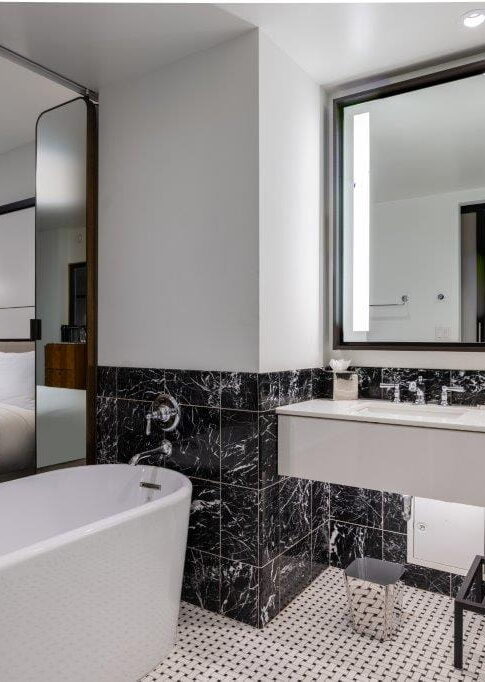 Modern hotel bathroom with a white bathtub, black marble walls, large mirror, and a view into a bedroom with a bed and lamp.