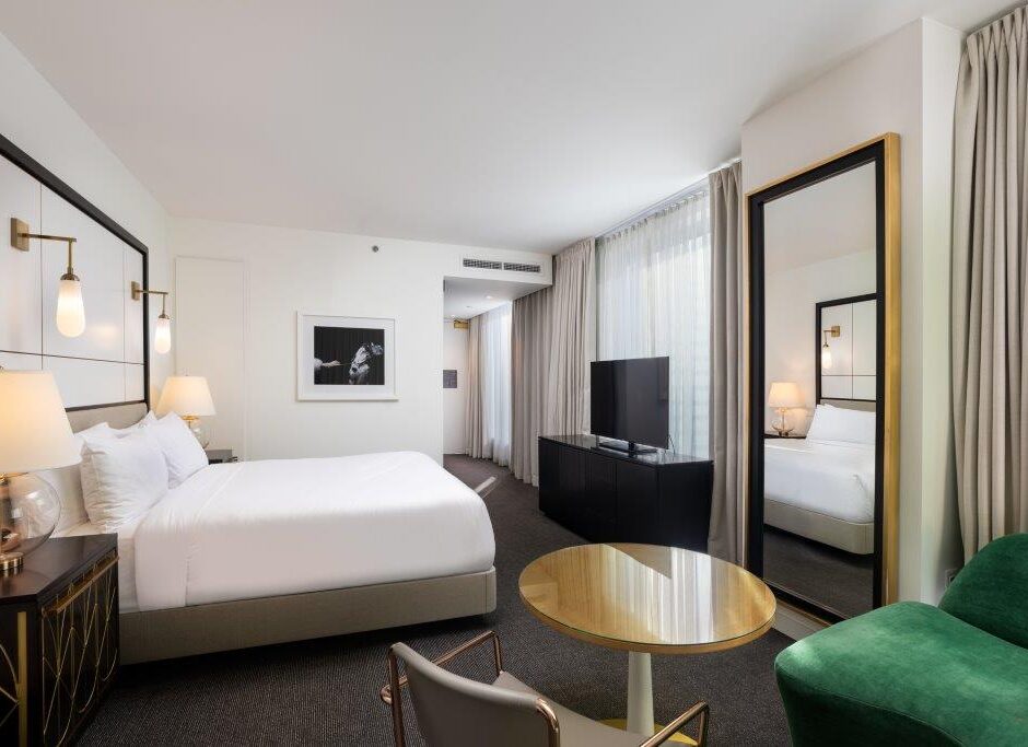 Modern hotel room with a large bed, TV, seating area, round table, green chair, and large mirror near a window with sheer curtains.