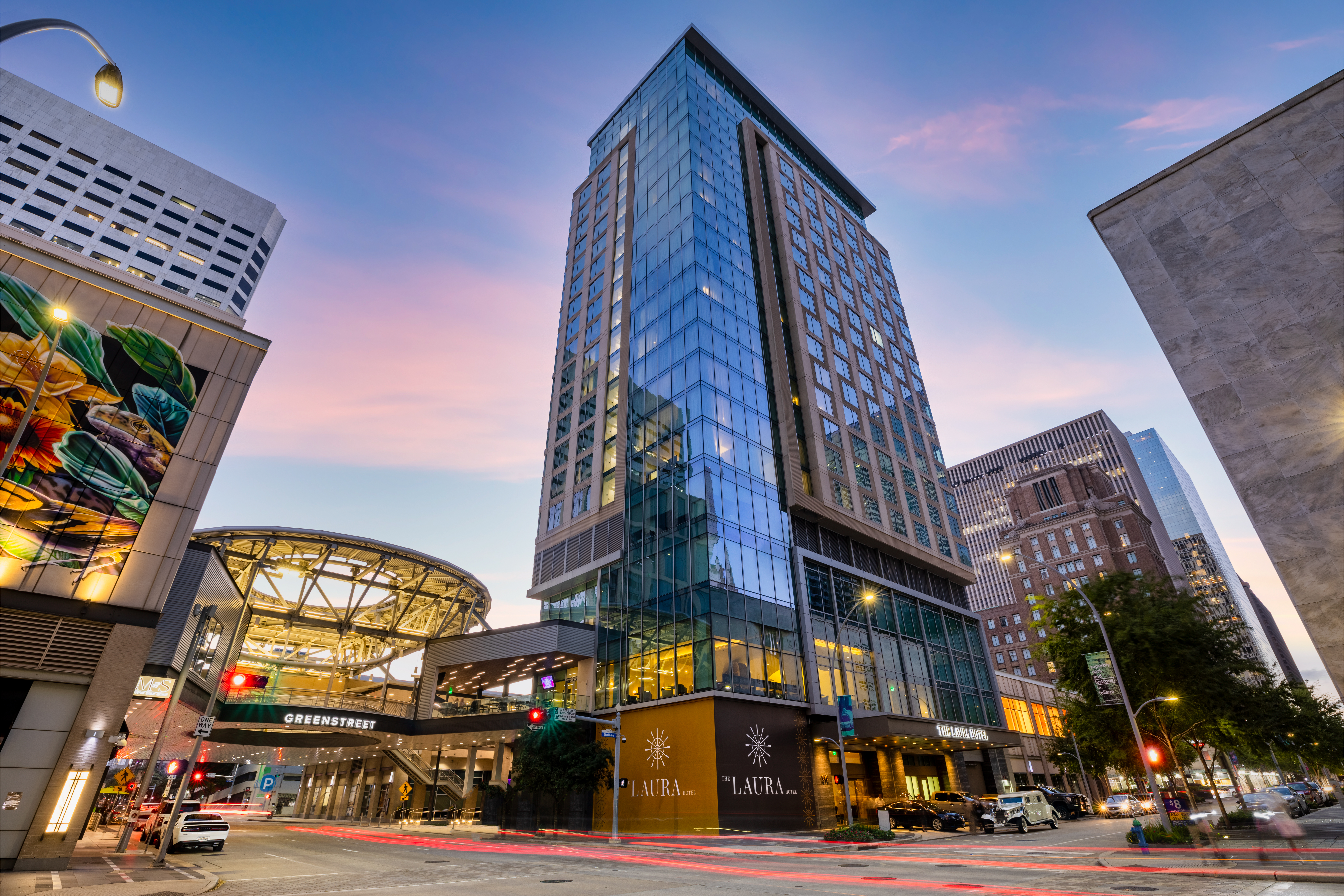 Exterior of The Laura Hotel in downtown Houston at dusk, featuring its glass high-rise facade, illuminated entrance and surrounding city buildings