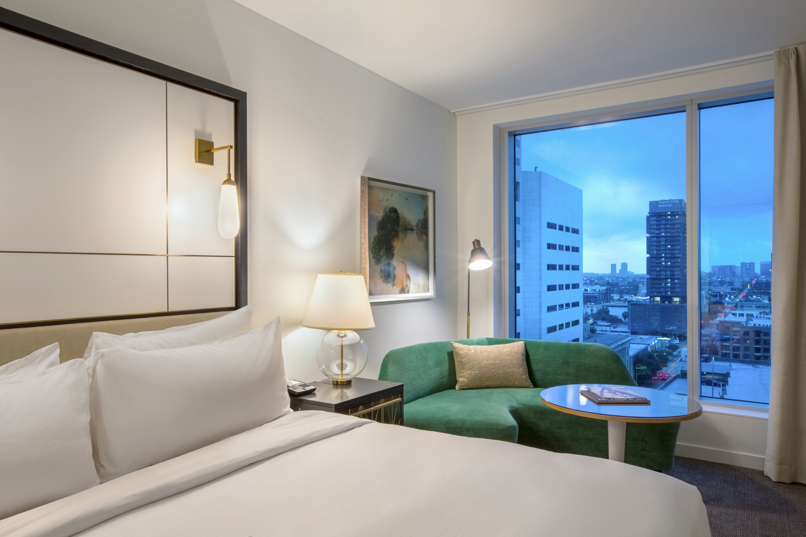 Modern hotel room with a white bed, green sofa, lamps, artwork, and a large window showing city buildings at dusk.