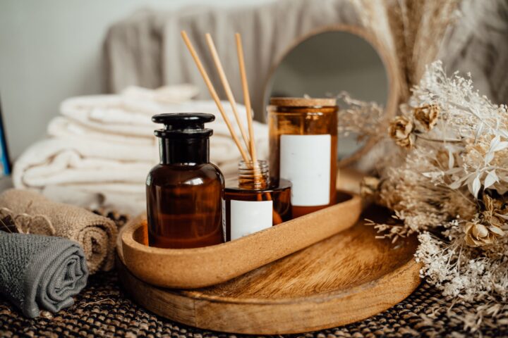 Brown glass bottles and reed diffuser on a wooden tray, with towels and dried flowers nearby, creating a cozy, spa-like atmosphere.