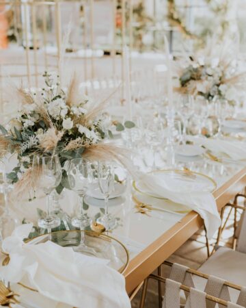 Elegant table set for an event with white flowers, glassware, white napkins, and gold accents on a mirrored surface.