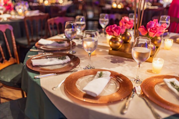 Elegant round table set with plates, napkins, wine glasses, and pink rose centerpieces in a softly lit event setting.