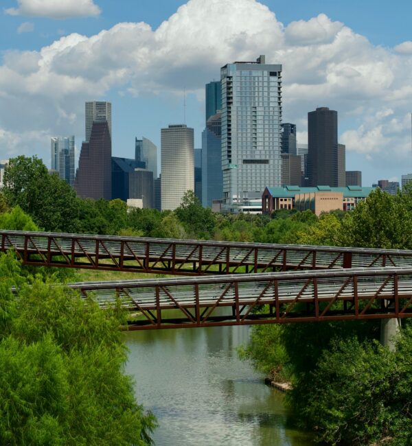 A metal bridge spans over a river with lush green trees, and city skyscrapers rise in the background under a blue sky with clouds.