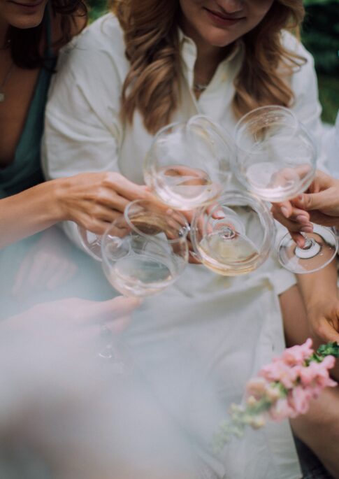 Four women sit closely and clink wine glasses in a celebratory toast, with soft focus and a relaxed, outdoor setting.