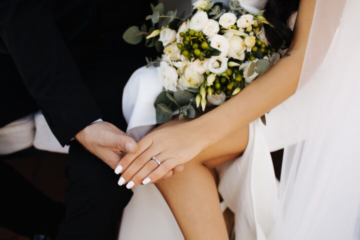 Bride and groom holding hands, bride wearing a white dress and engagement ring, holding a bouquet of white flowers and greenery.