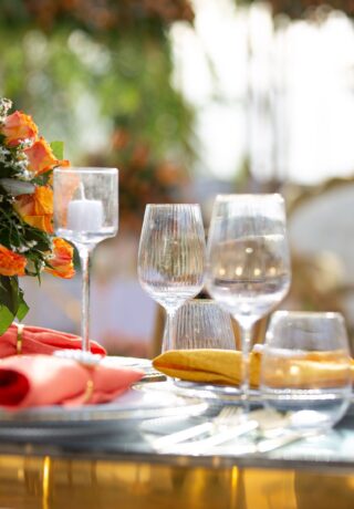 Elegant outdoor table with orange flowers, glassware, and plates. Napkins are neatly folded in red and yellow on a reflective surface.