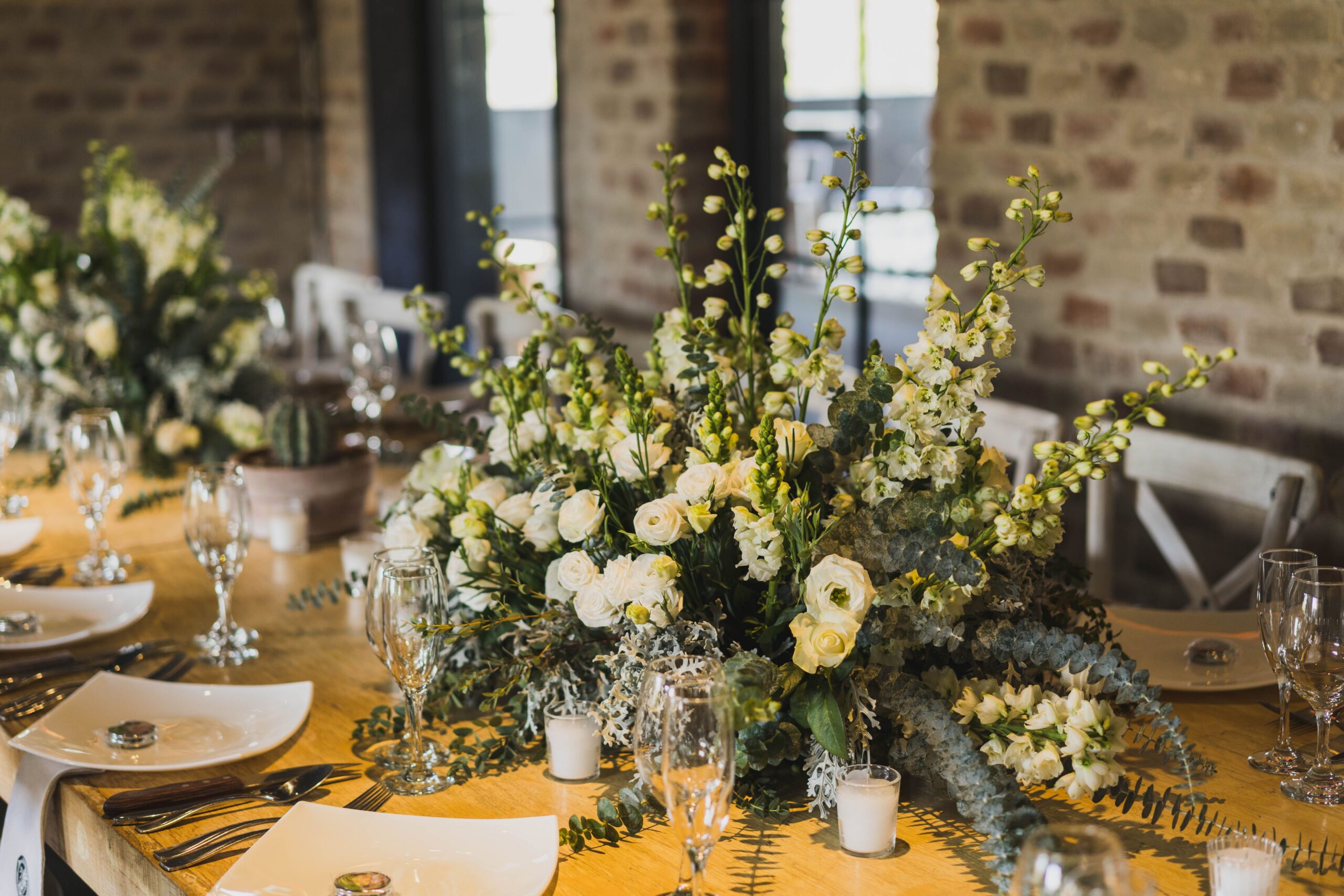 Elegant floral centerpiece with white and yellow flowers on a wooden table set for a formal meal, with plates, glasses, and candles.