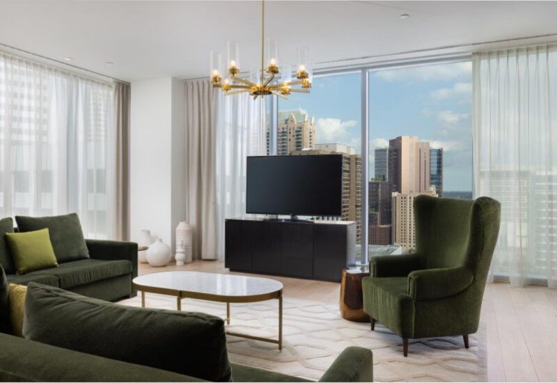 Modern living room with green sofas, a TV, chandelier, coffee table, and large windows showing a city skyline with tall buildings.