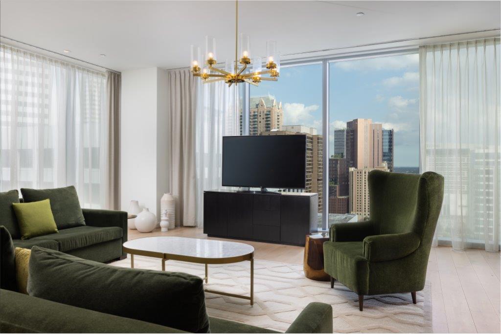 Modern living room with green sofas, a TV, chandelier, coffee table, and large windows showing a city skyline with tall buildings.