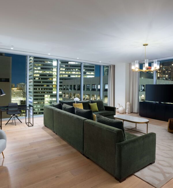Modern living room with large windows, city night view, green sectional sofa, armchair, round coffee table, TV, and a small desk by the window.