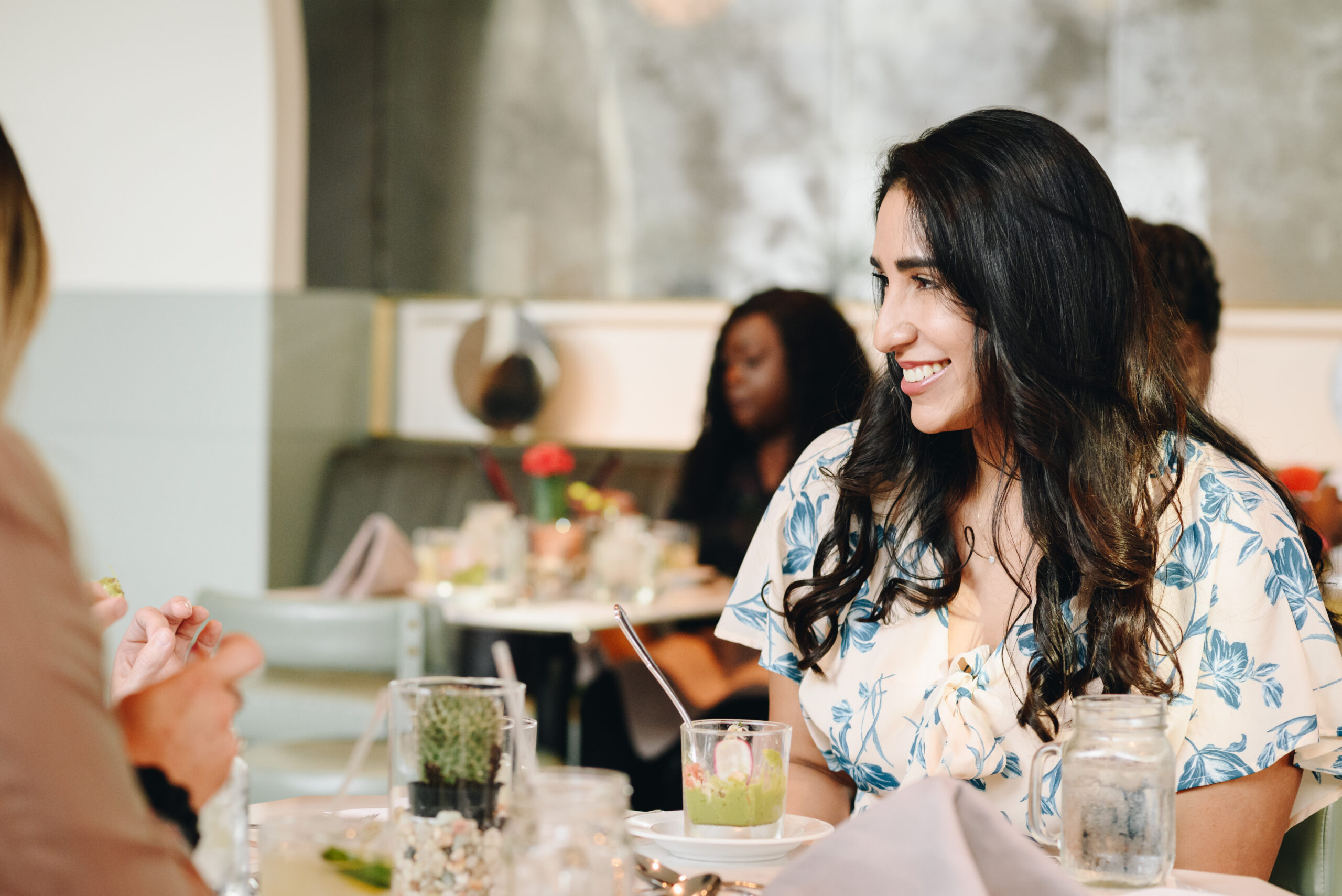 Woman in floral dress smiles while dining with others at a restaurant; blurred people and table settings are visible in the background.