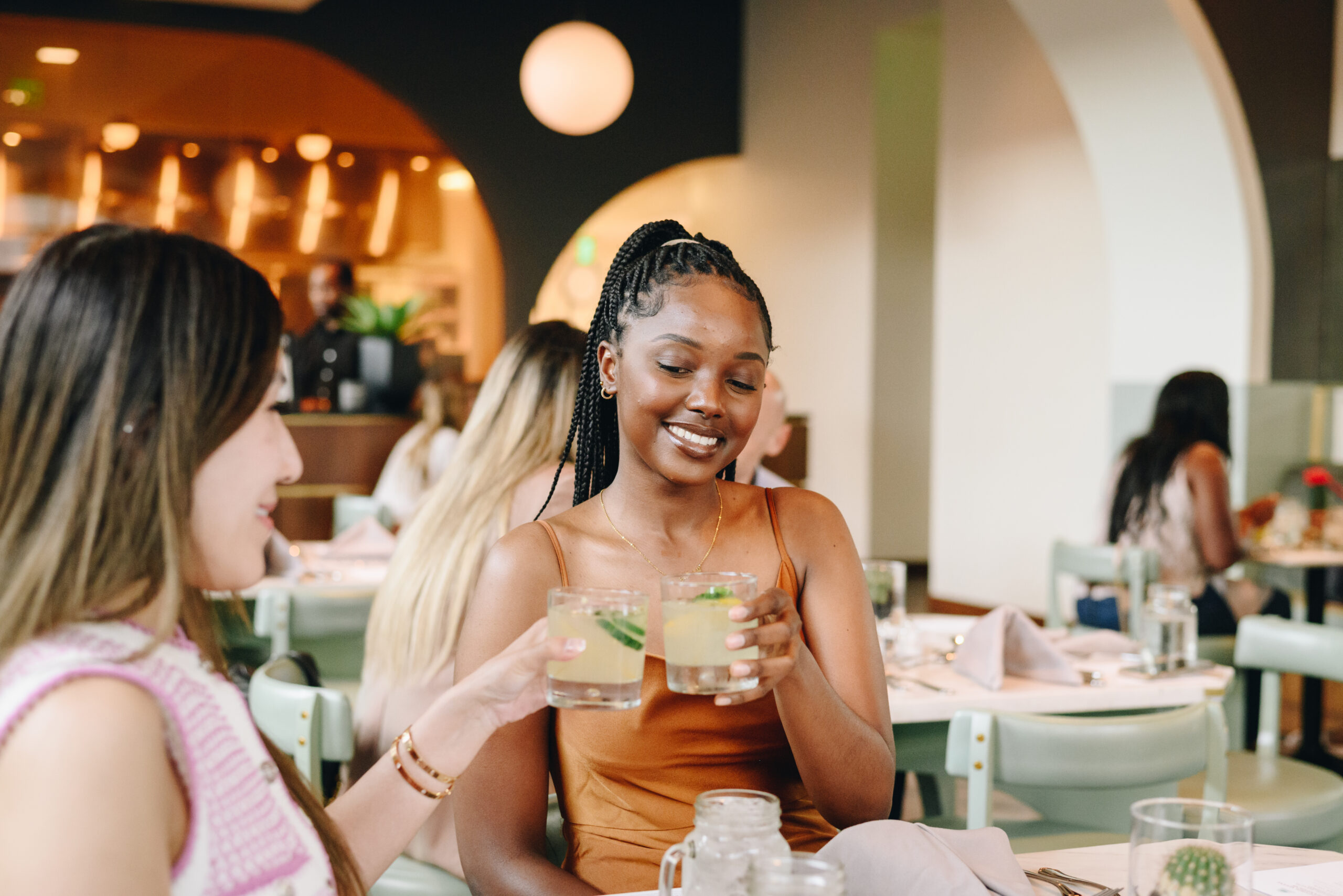 Two women sit at a restaurant table, smiling and clinking glasses with drinks, surrounded by other diners in a modern, well-lit setting.