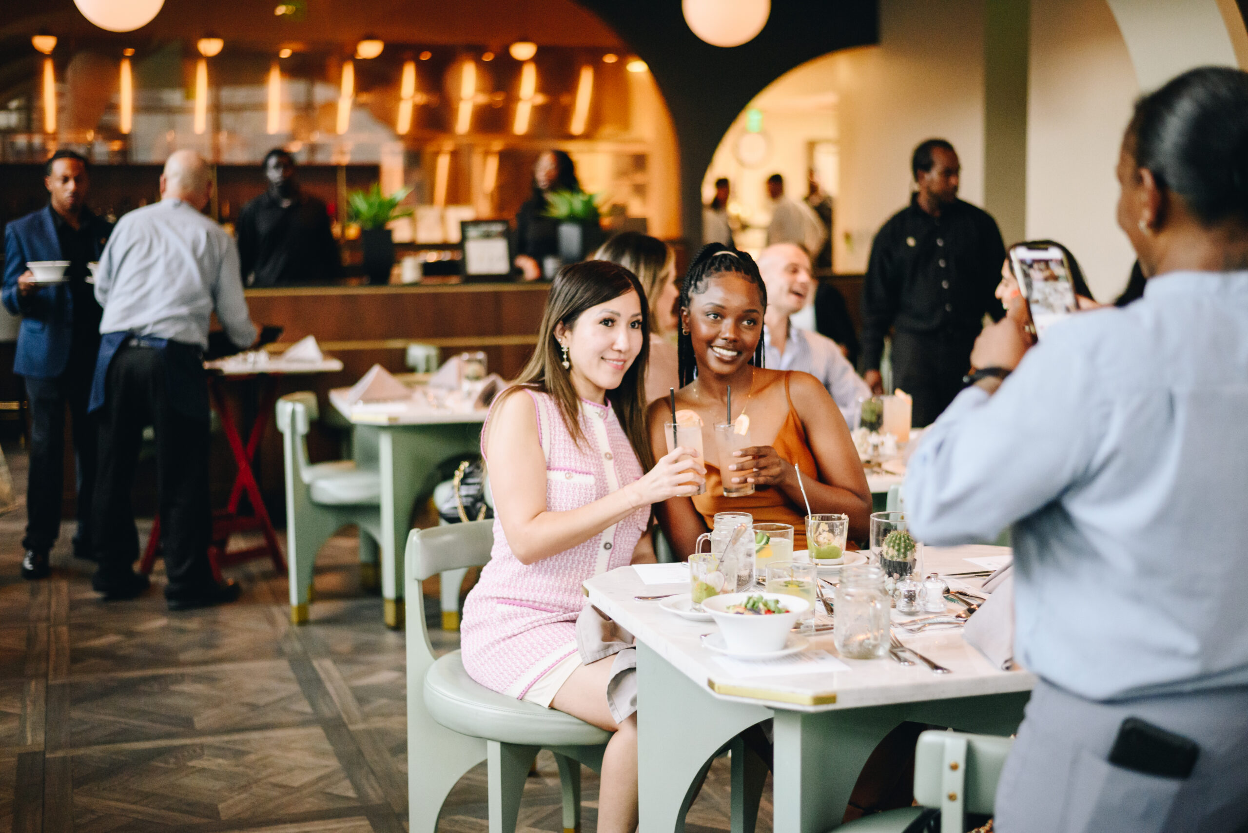 Two women sitting at a restaurant table smile and toast drinks while someone takes their photo; people and staff are in the background.