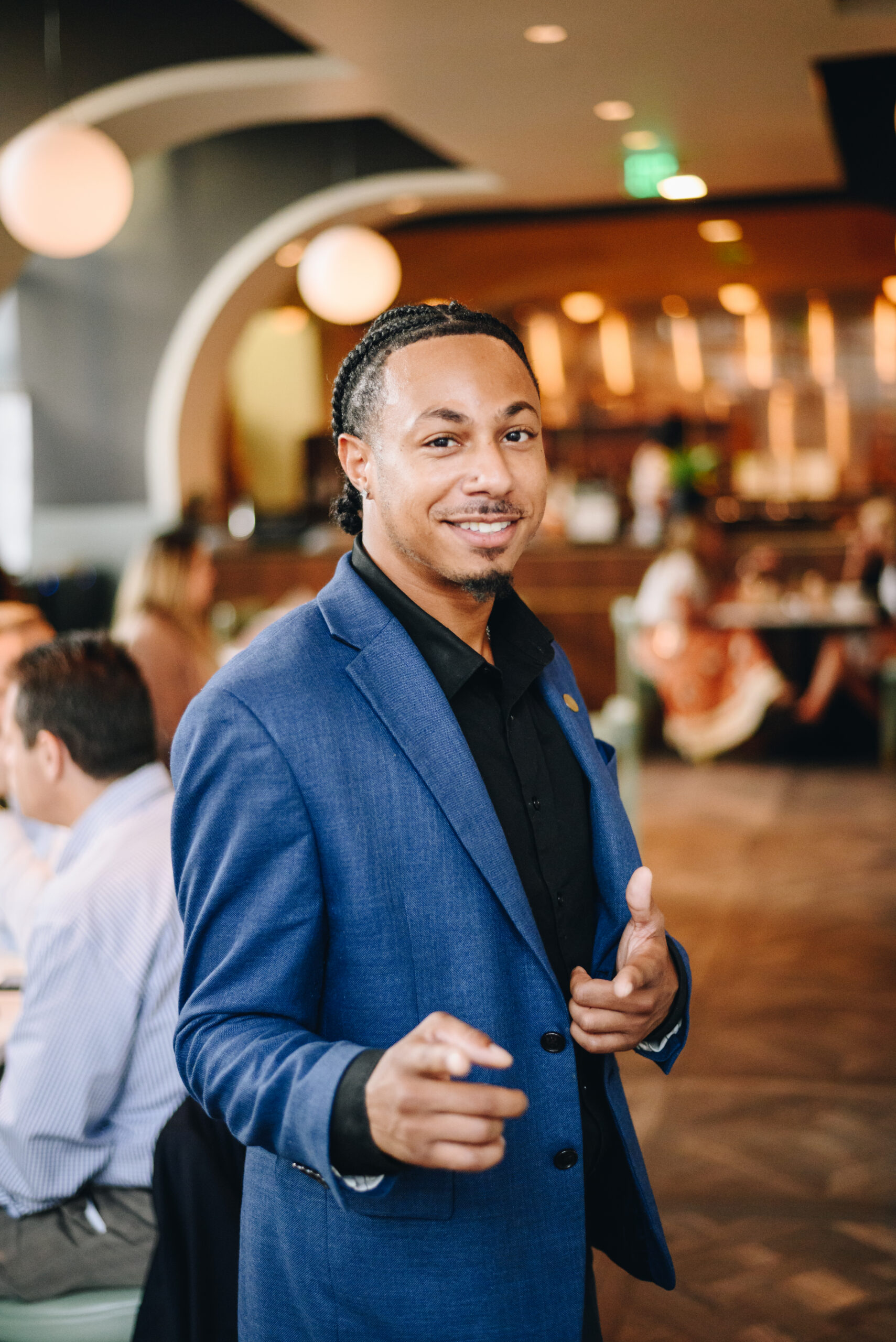 Man in blue suit smiling and pointing at the camera in a restaurant with people blurred in the background.