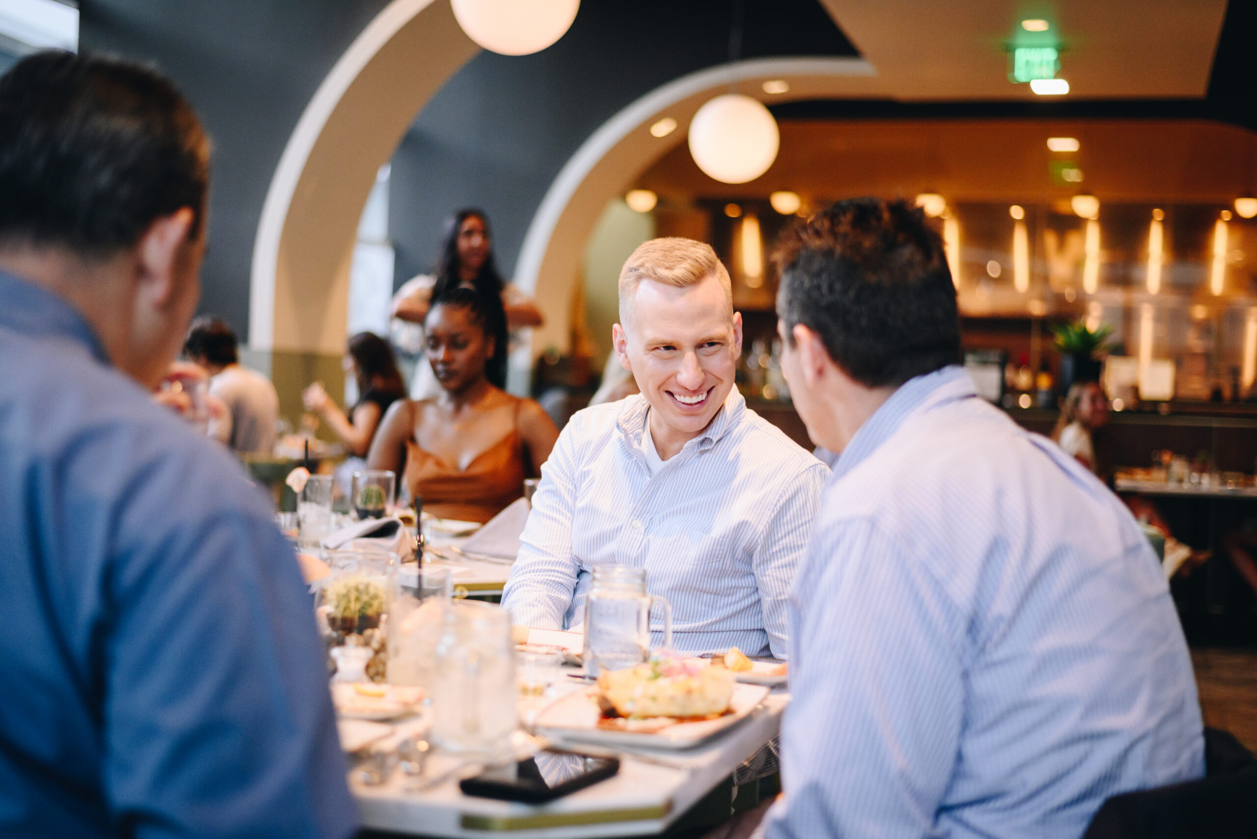 Two men are smiling and talking at a restaurant table, while others dine and chat around them in a bright, modern setting.