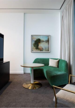 Modern hotel room with a green sofa, round table, gold chair, TV divider, wall art, and floor-to-ceiling window with city view.