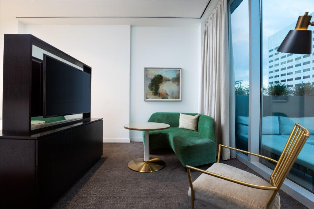 Modern hotel room with a green sofa, round table, gold chair, TV divider, wall art, and floor-to-ceiling window with city view.