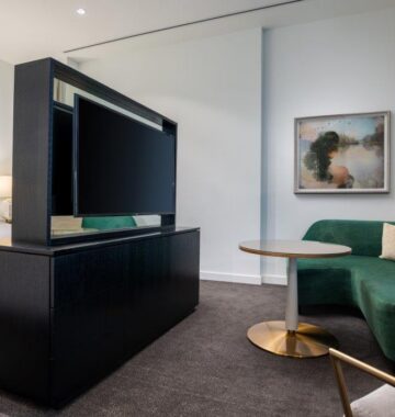 Modern hotel room with a bed, divider-mounted TV, green couch, round table, and artwork on a white wall.