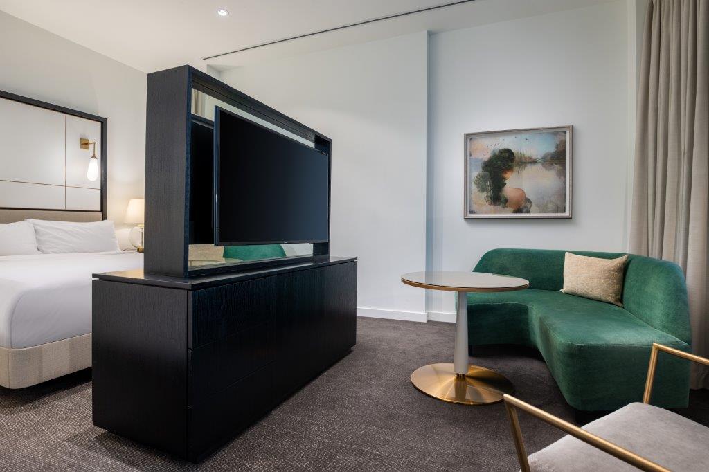 Modern hotel room with a bed, divider-mounted TV, green couch, round table, and artwork on a white wall.