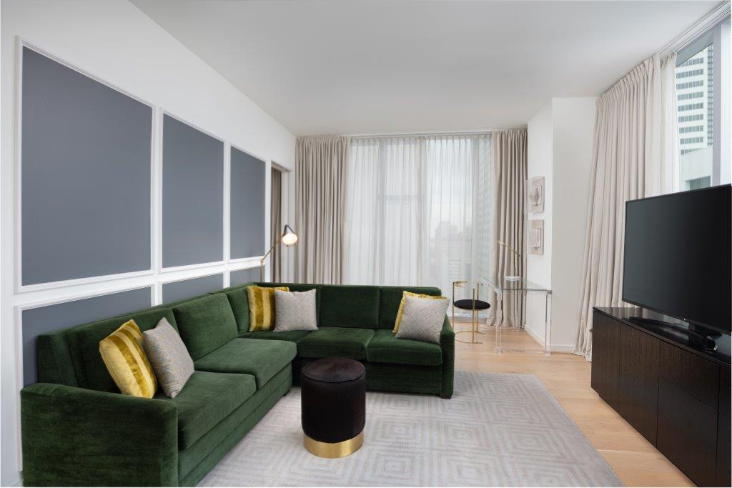 Modern living room with a green sectional sofa, gold-accented pillows, TV, black cabinet, large windows, neutral walls, and soft lighting.