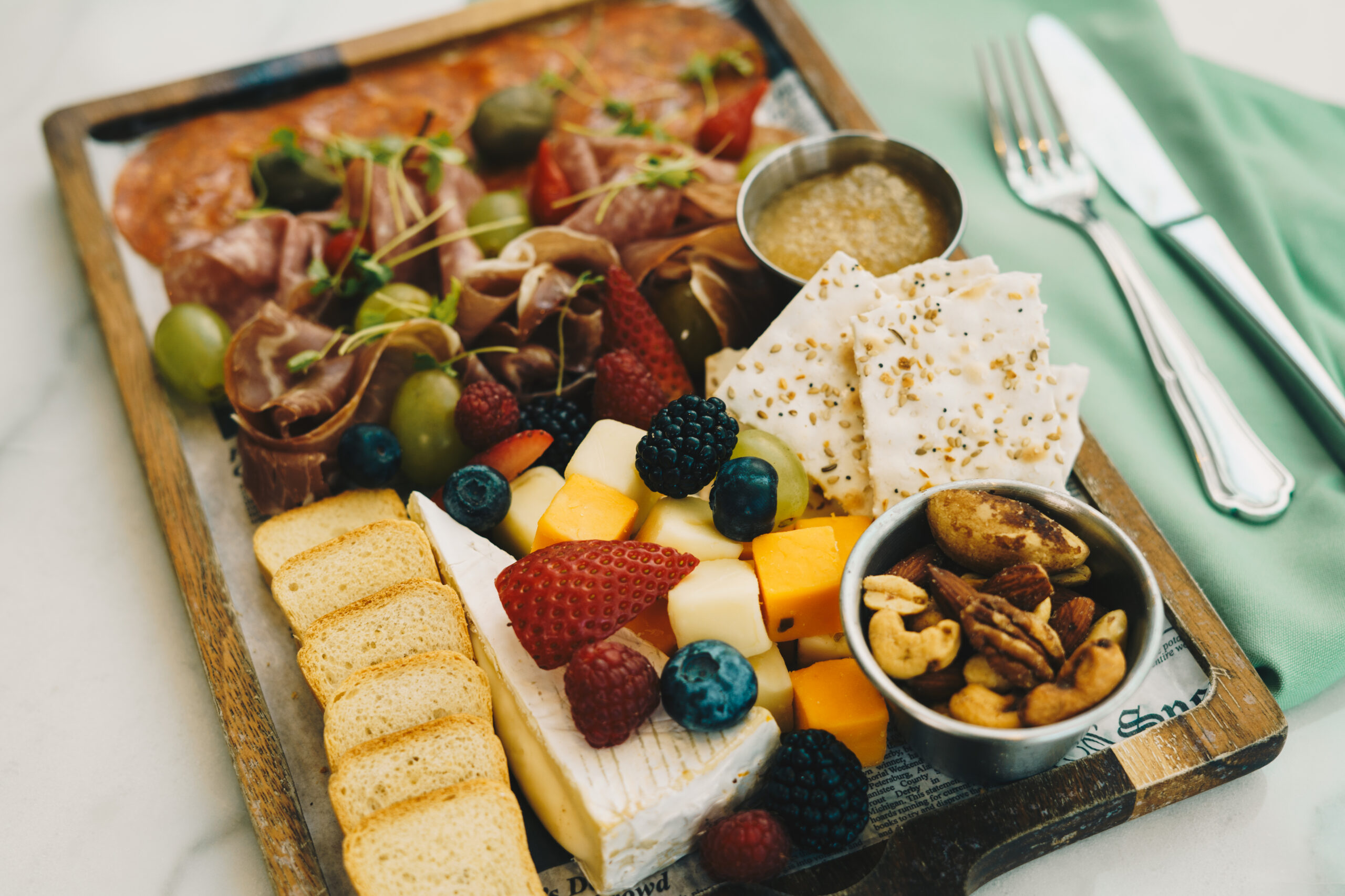 A charcuterie board with meats, cheeses, crackers, berries, nuts, olives, and honey, placed next to a fork and knife on a green napkin.