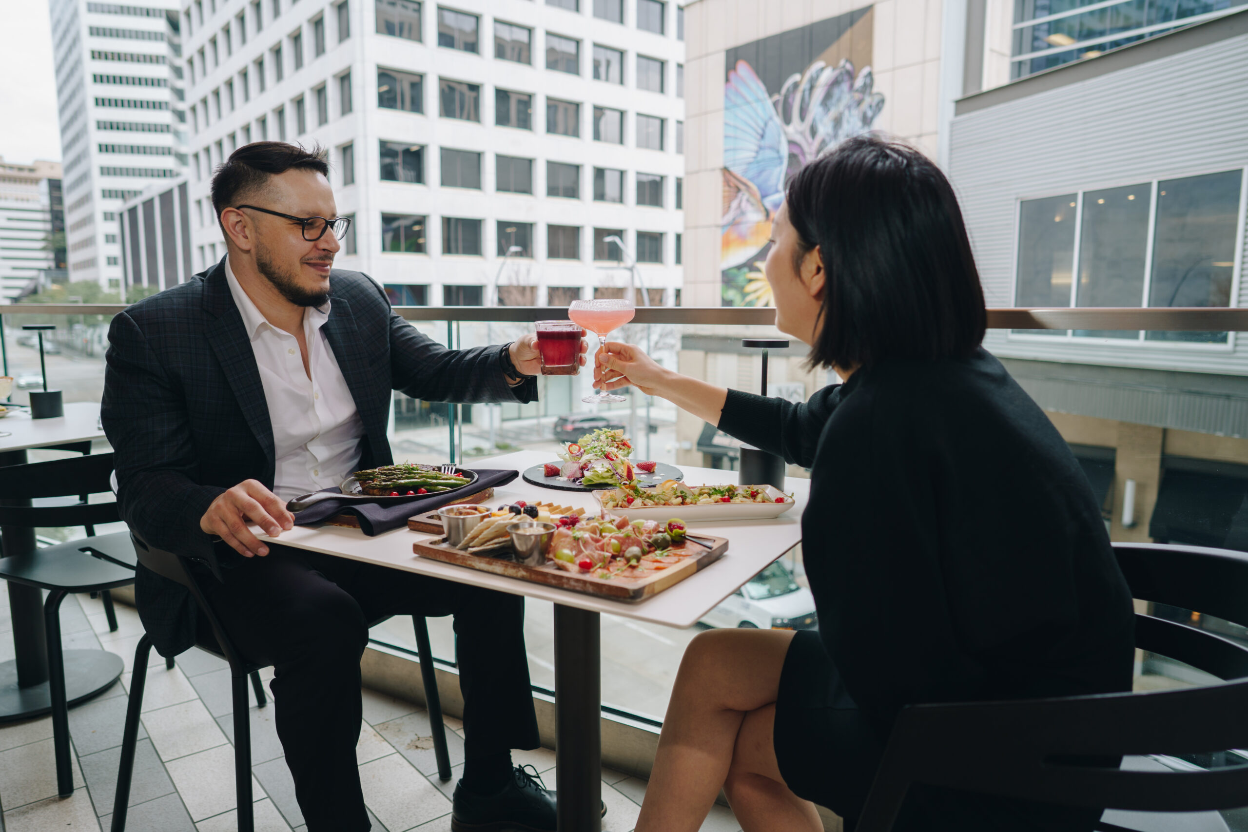 Two people sit at an outdoor table in a city, toasting drinks and enjoying a meal with flatbreads and salads.