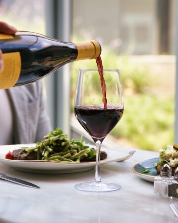 A person pours red wine into a glass at a restaurant table with plates of food and silverware.