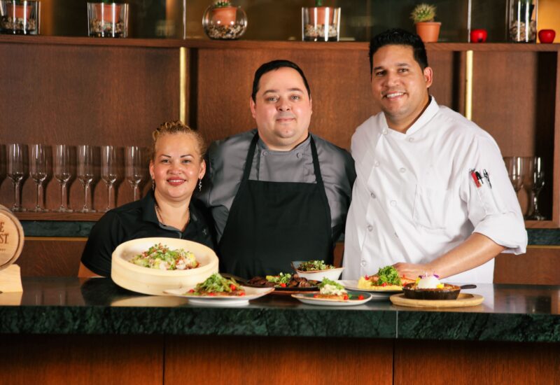 Three chefs stand behind a counter displaying plated food, smiling at the camera in a restaurant kitchen setting.