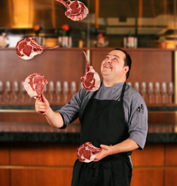 A chef in a kitchen juggles five large raw tomahawk steaks, smiling as he tosses them in the air.