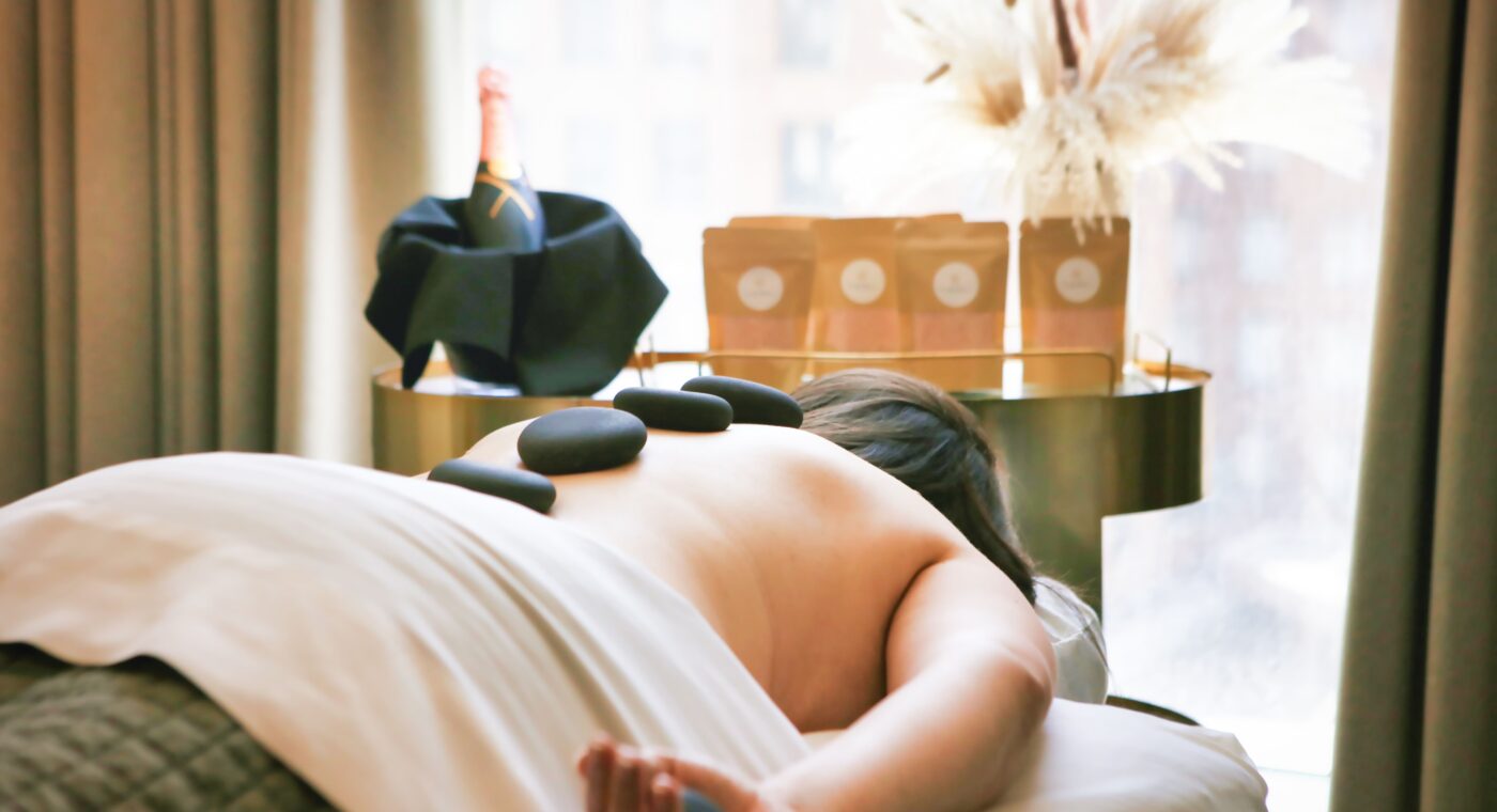 Person lying face down on a massage table with hot stones on their back, near a window and a table holding champagne and decorative items.