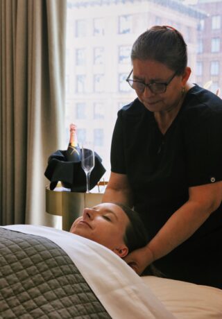 A woman receives a neck massage from a therapist in a spa room with a champagne bottle and glass on a table in the background.