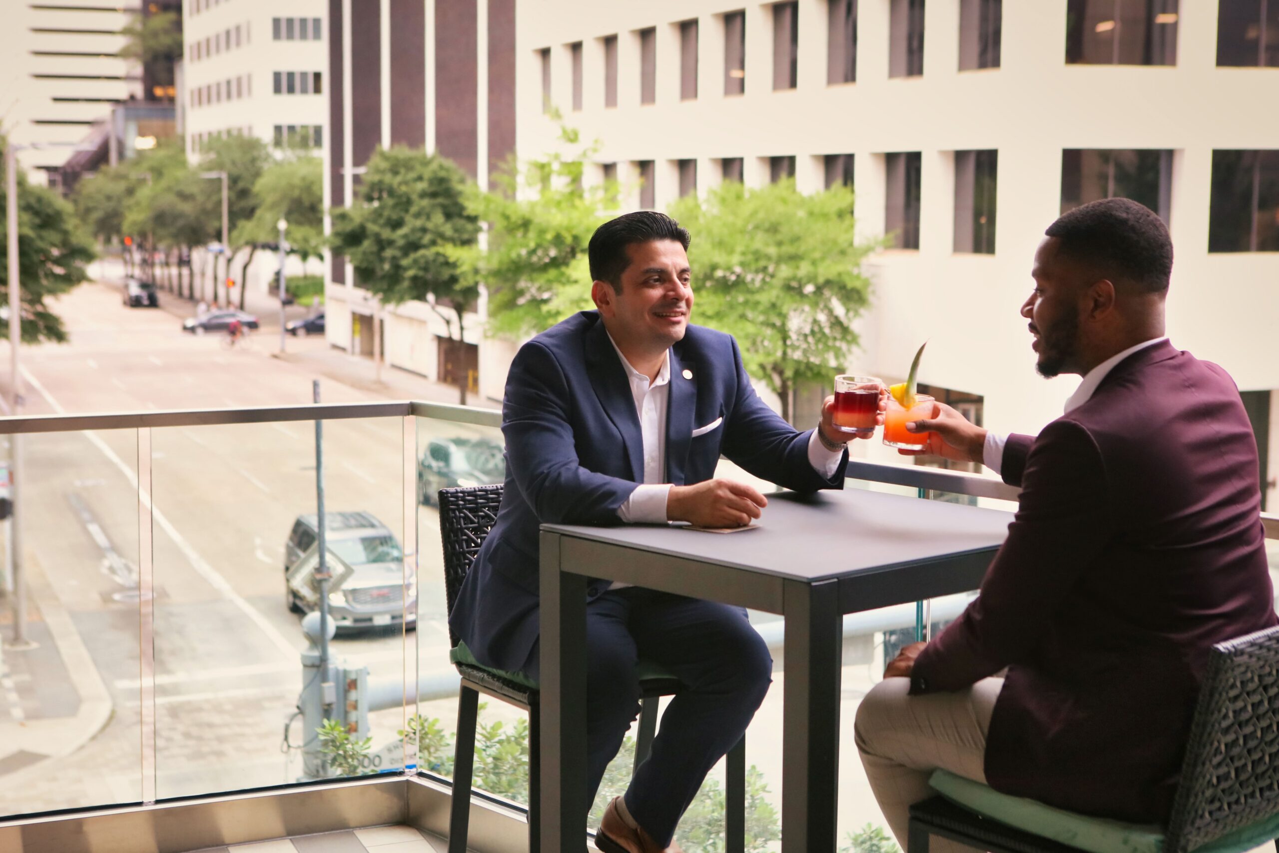 Two men in suits sit at a balcony table in a city, smiling and clinking glasses with drinks.