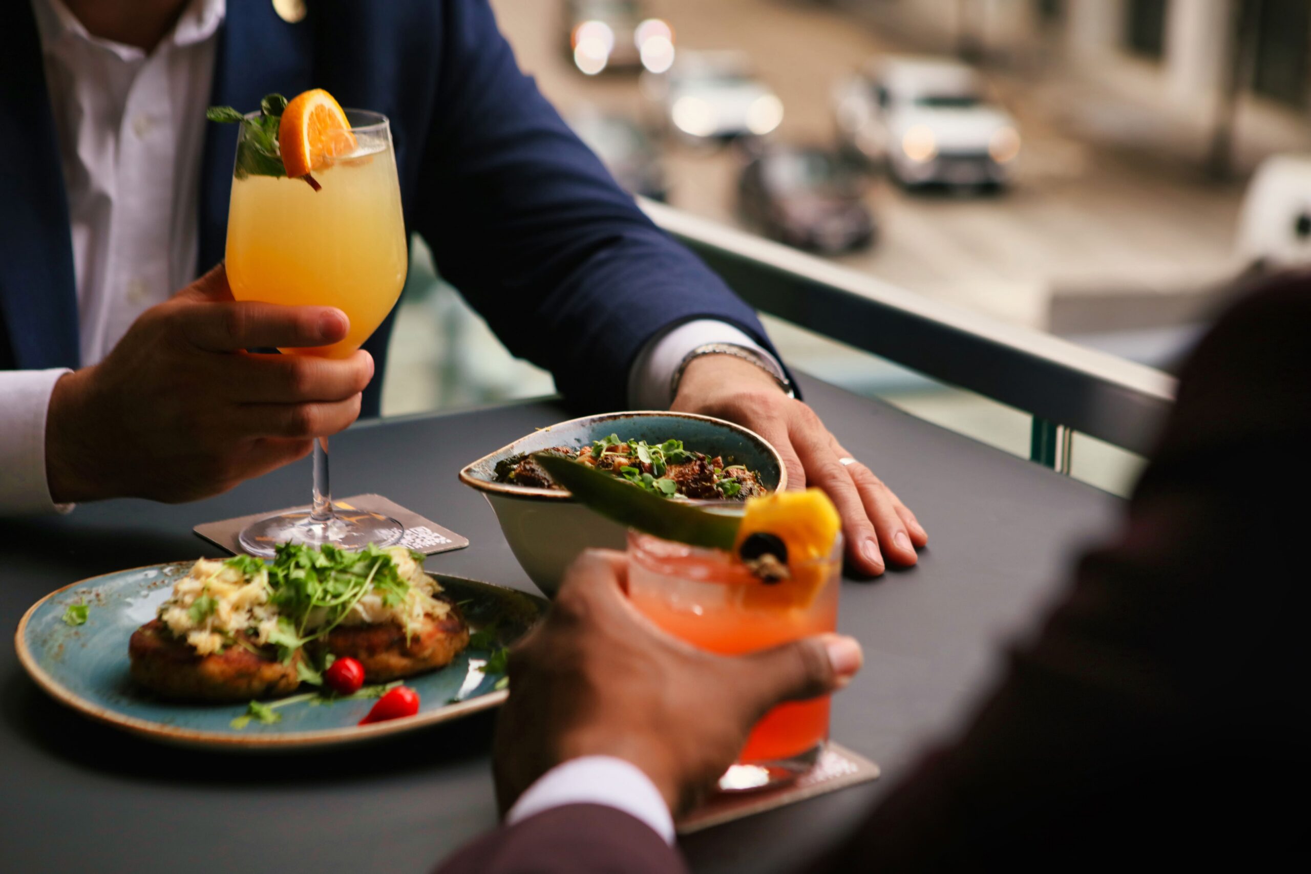 Two people in suits holding cocktails at an outdoor table with plates of food, sharing a meal in a city setting.