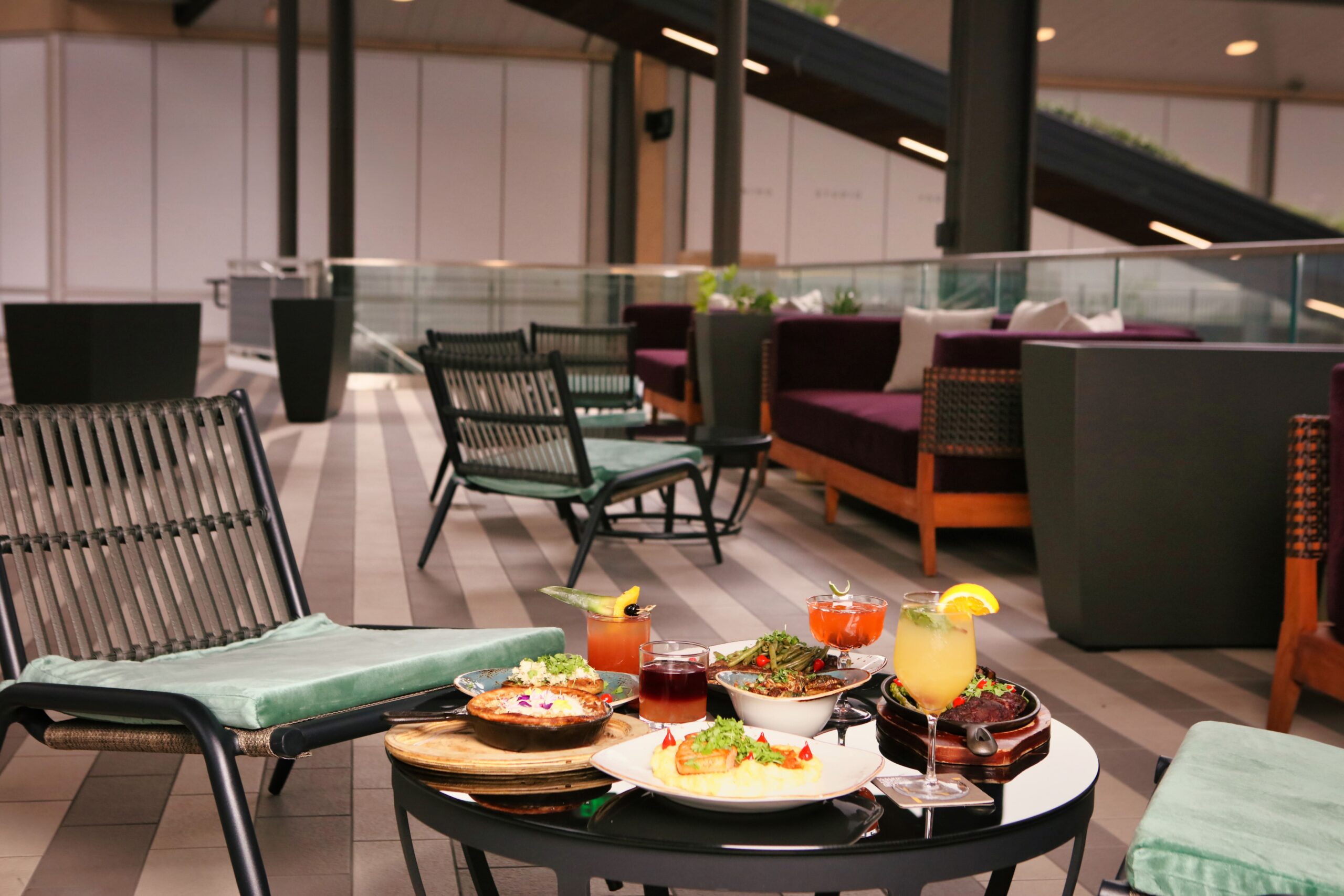 Outdoor lounge area with chairs and a small table set with colorful drinks and plates of food, surrounded by modern seating and plants.