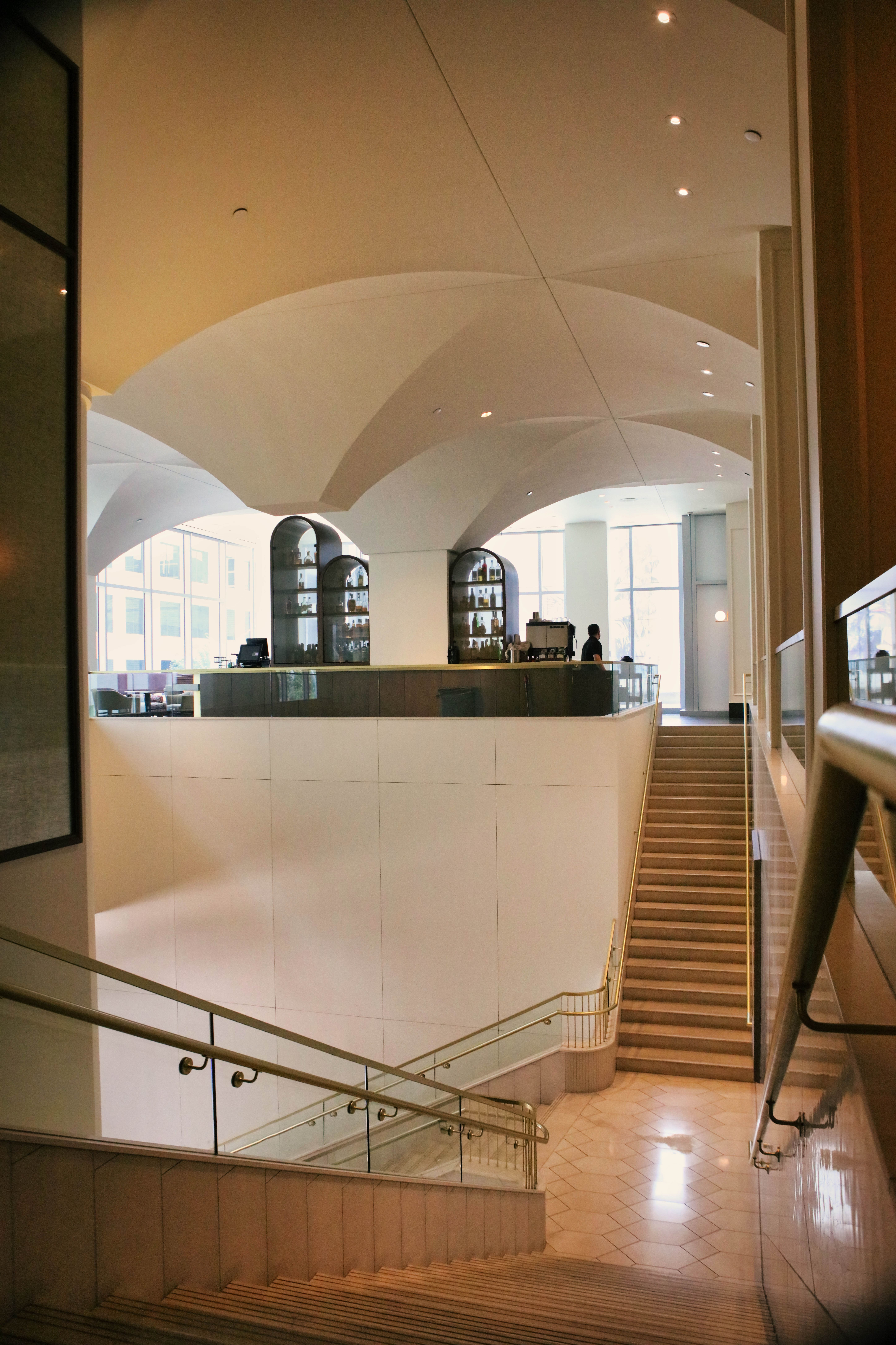 A wide, light-filled staircase leads up to an elegant bar area featuring arched ceilings, glass railings, and shelves filled with liquor bottles.
