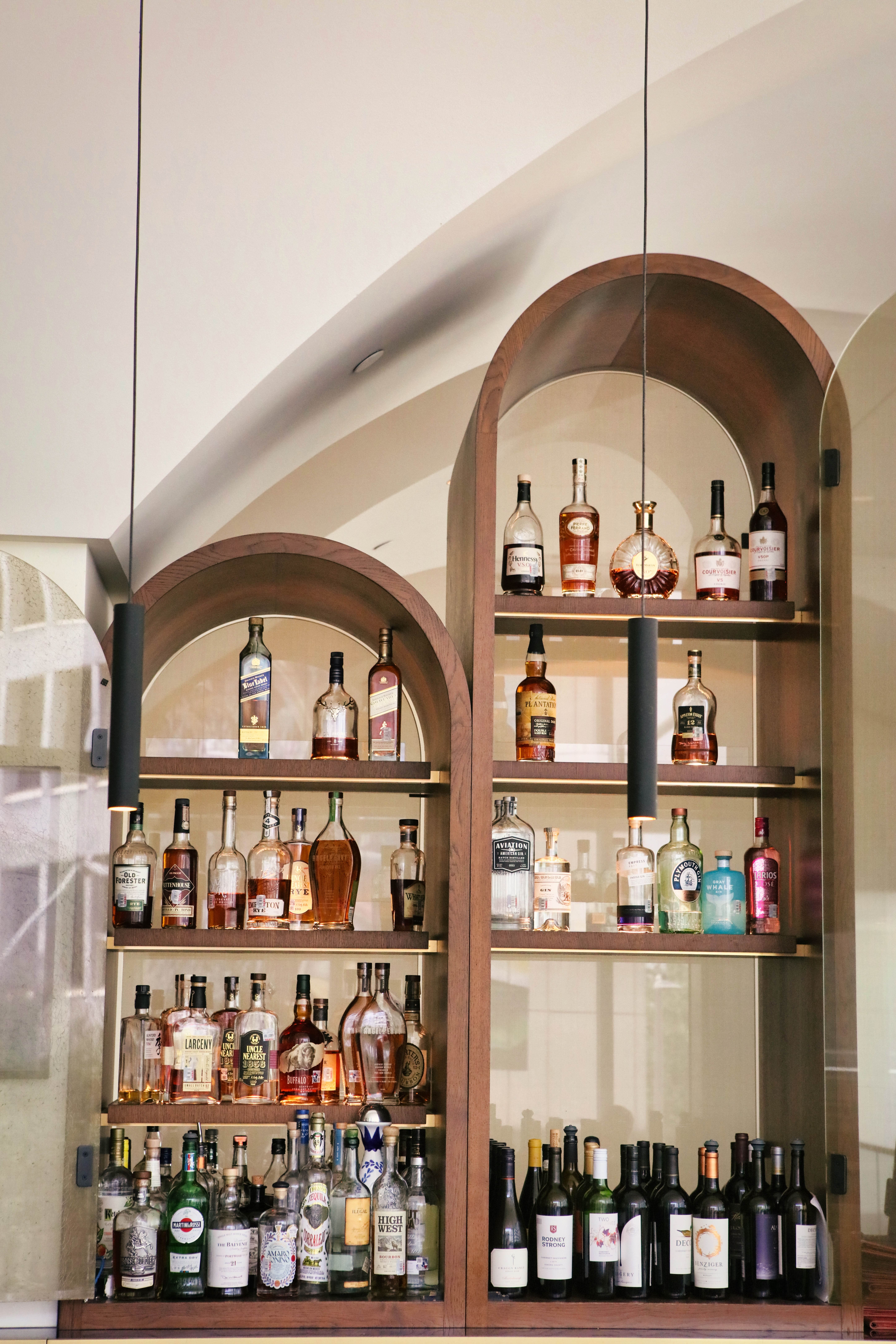 Two arched wooden shelving units filled with various liquor and wine bottles, illuminated by black pendant lights hanging from the ceiling.