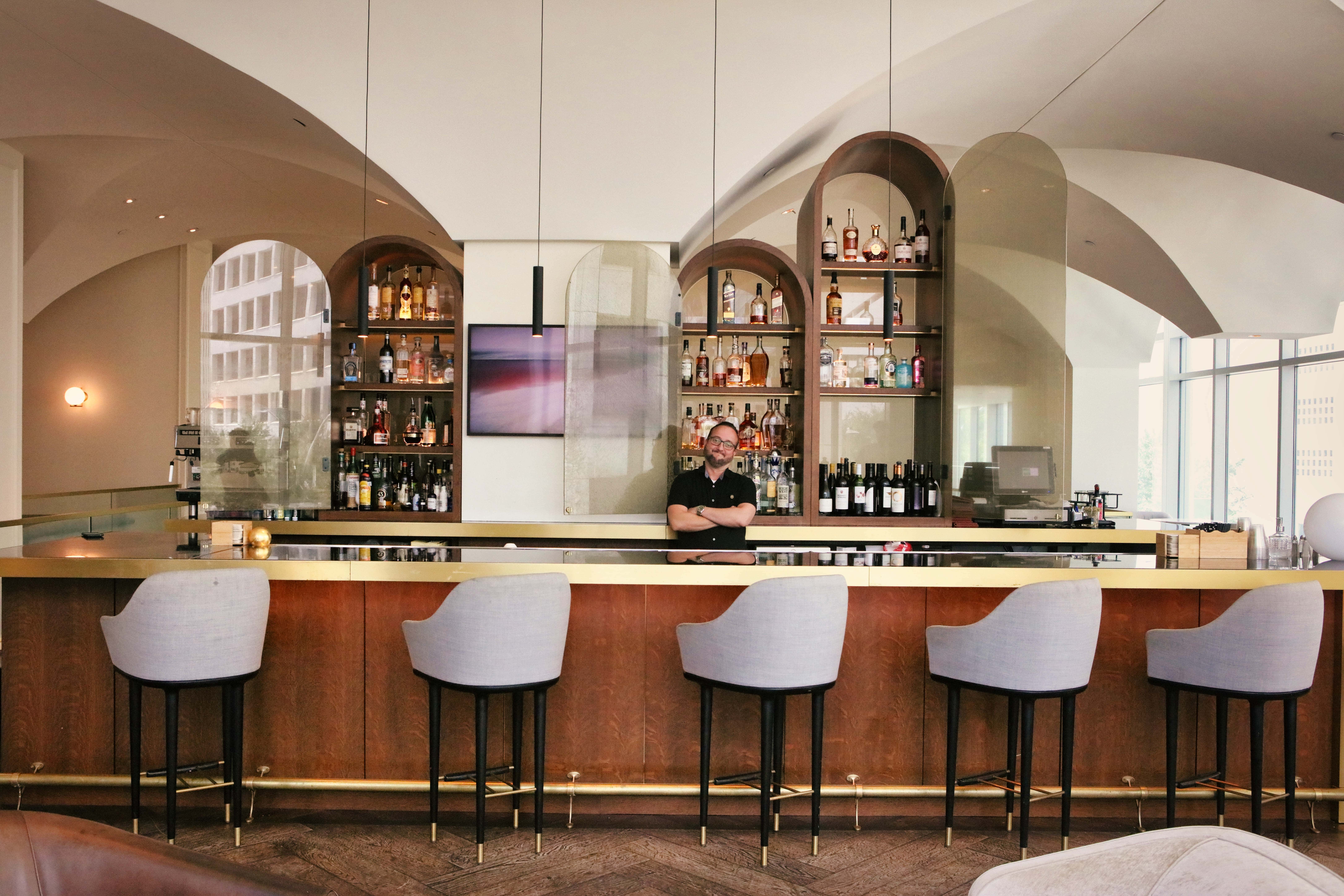 A bartender stands behind a modern bar with shelves of bottles, five empty gray chairs, and arched mirrors in a stylish, well-lit lounge.