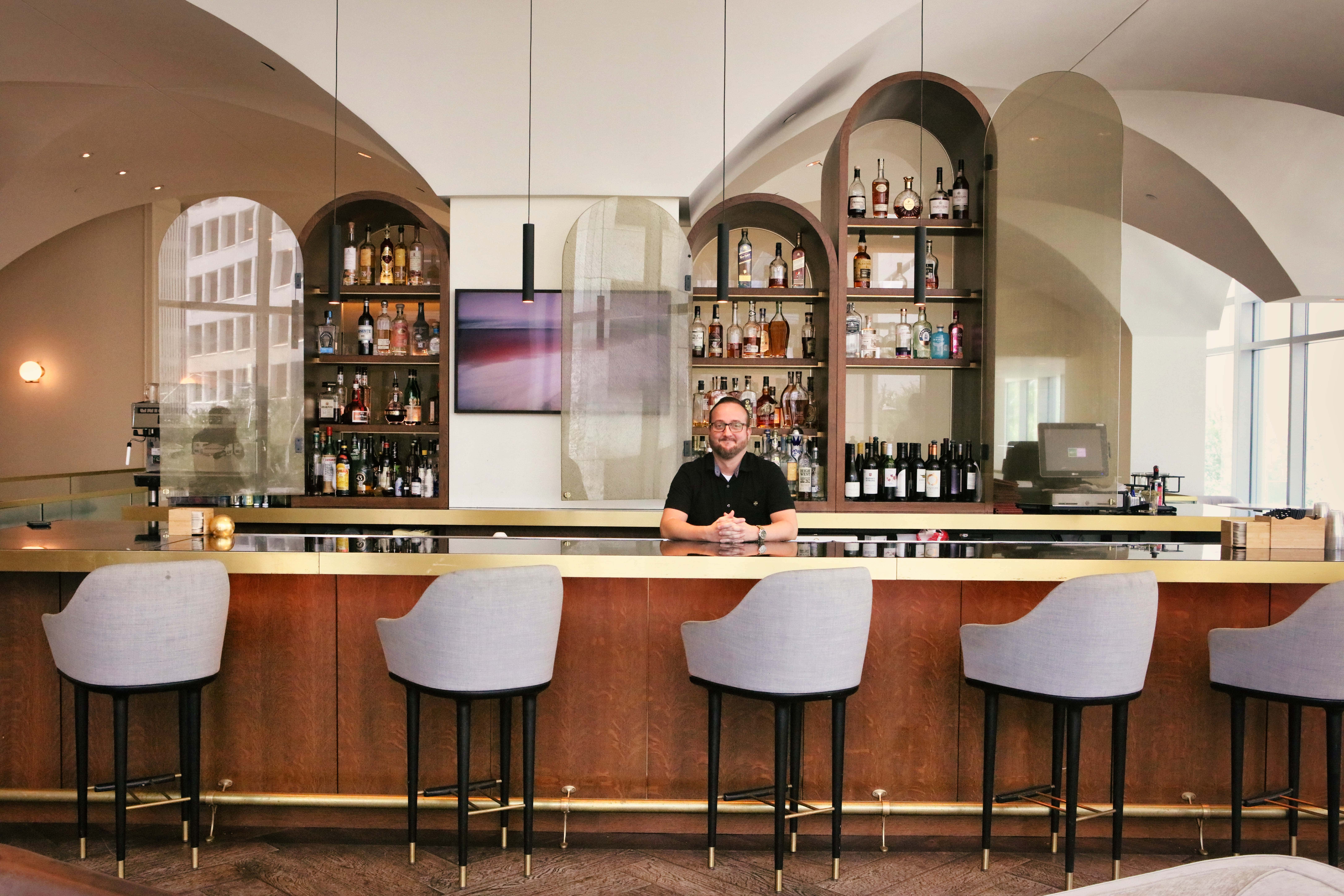 A bartender with glasses and a black shirt stands behind a modern, wood-paneled bar with gold trim and grey stools in a brightly lit, arched lounge.