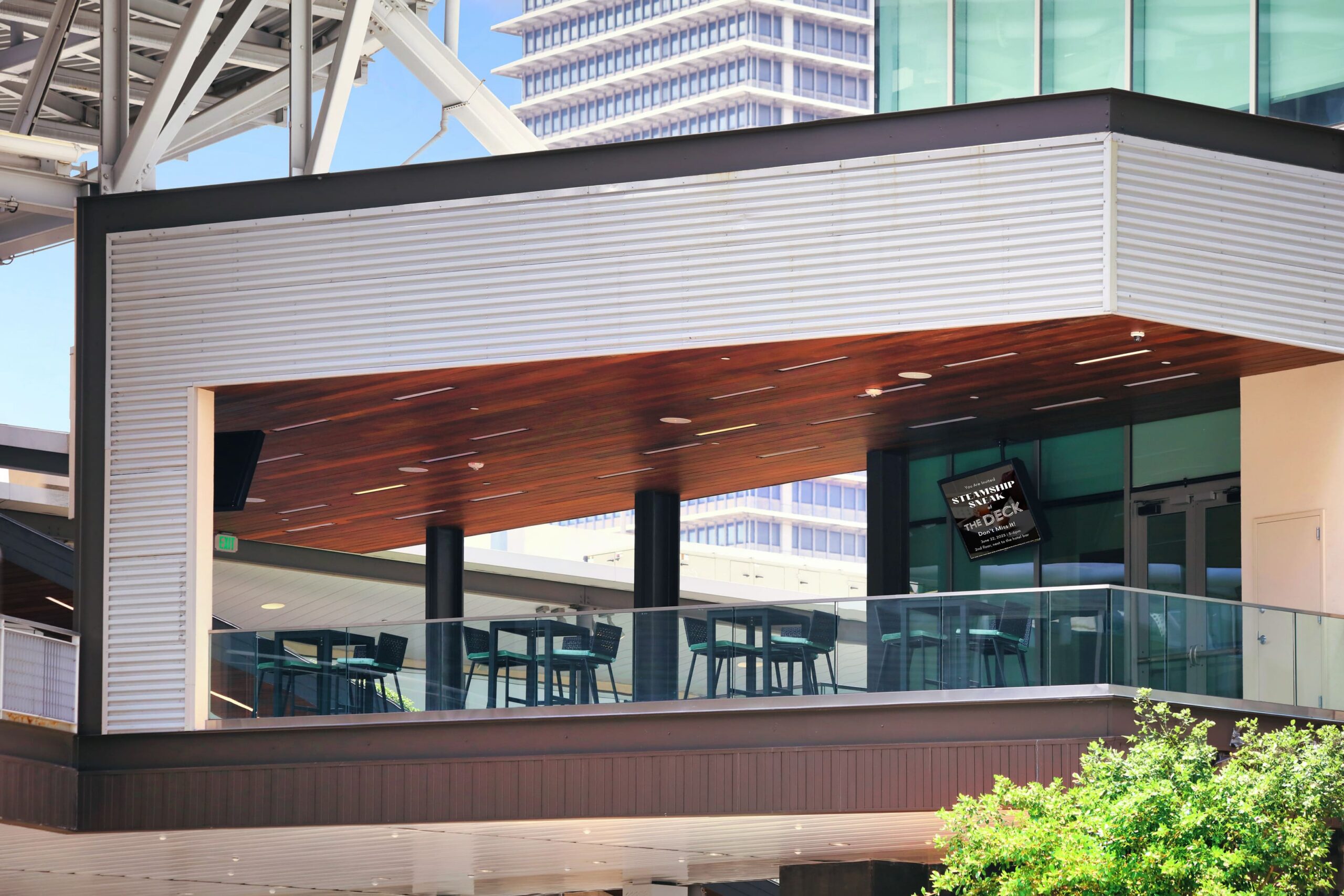 Modern outdoor balcony with tables and chairs, glass railing, and a partially open ceiling, with a city building visible in the background.