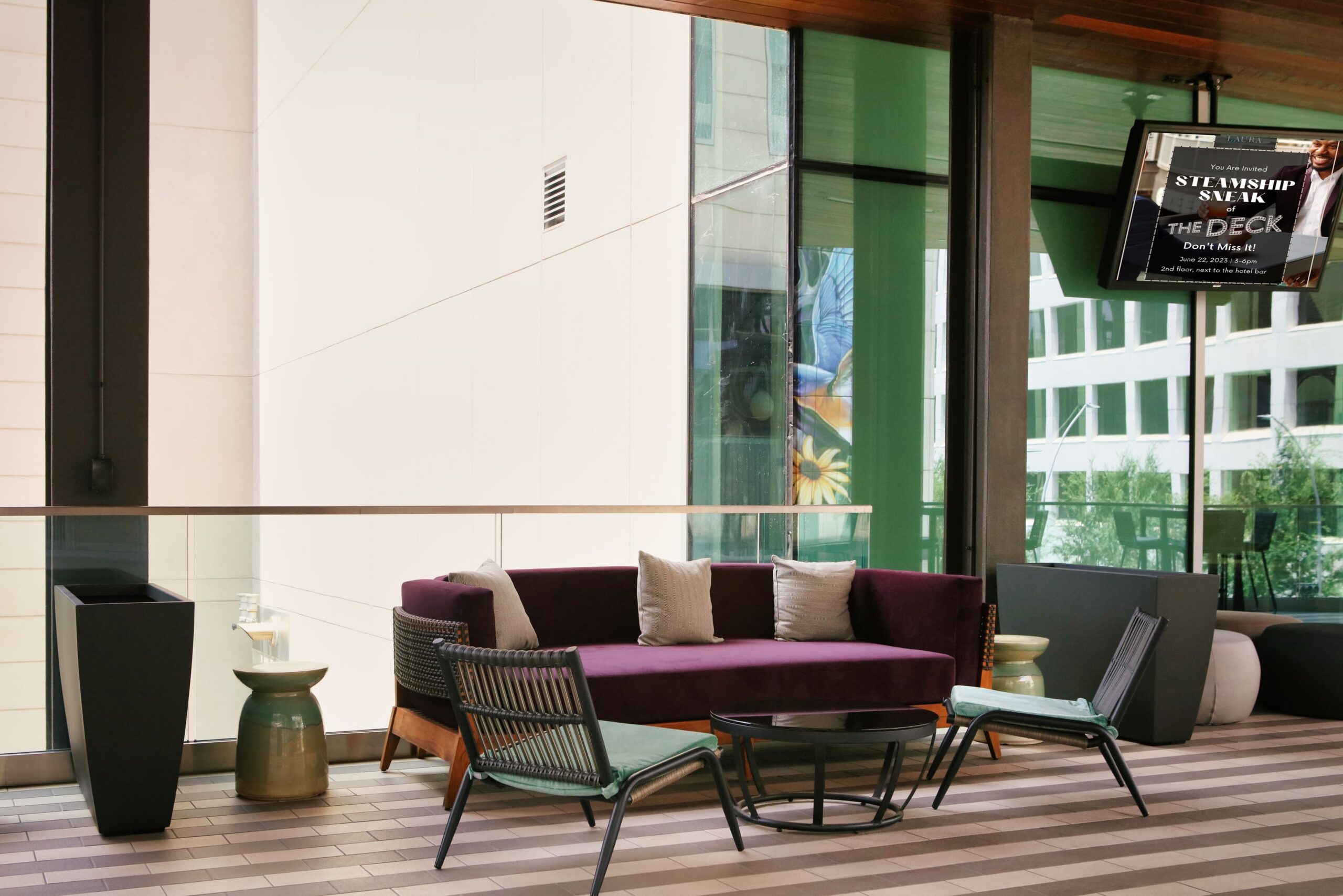 Modern outdoor lounge with a purple sofa, chairs, a small table, and a TV on the wall, with tall windows and city buildings visible outside.