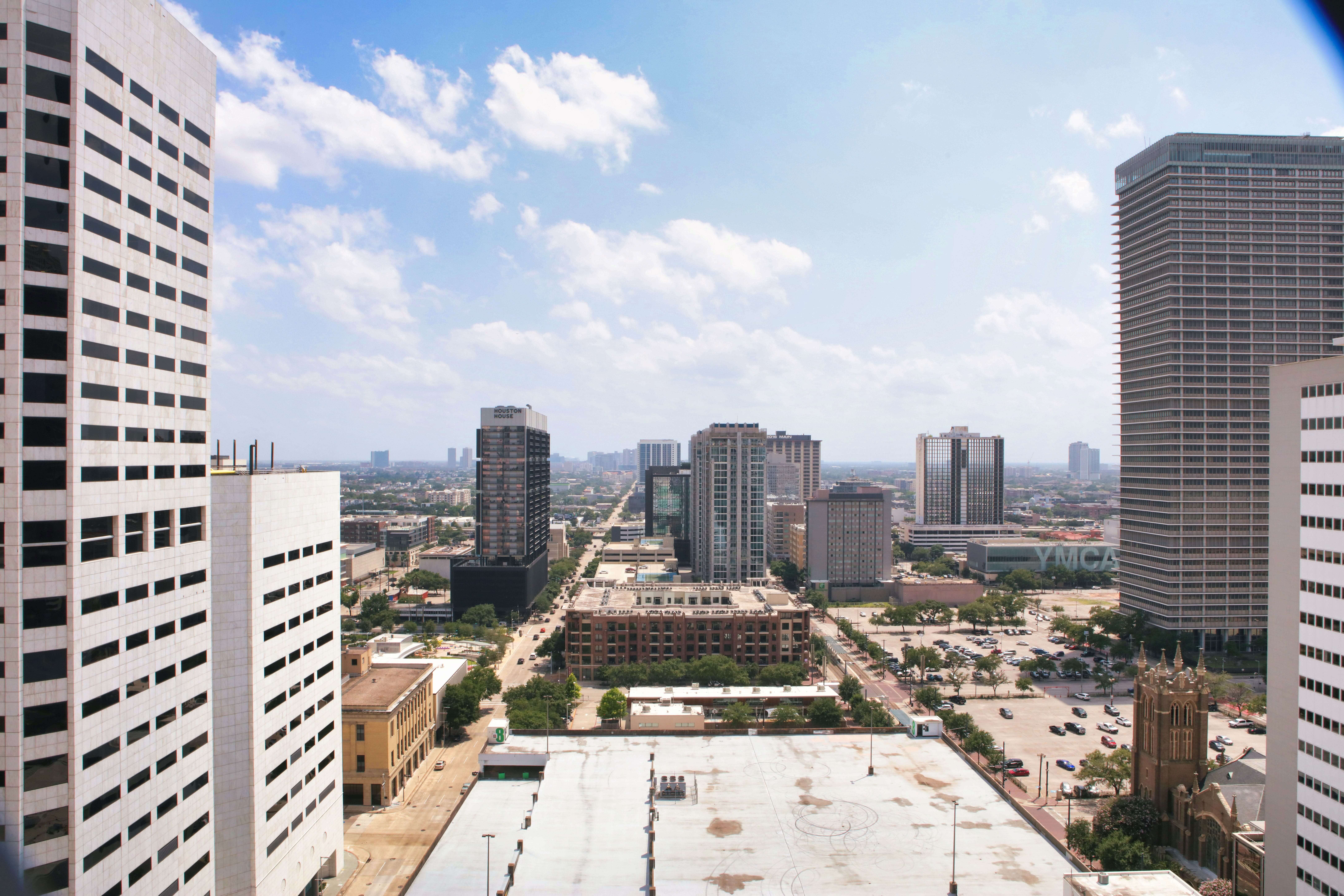 A cityscape view with tall modern buildings, blue sky, scattered clouds, and streets lined with cars and trees.