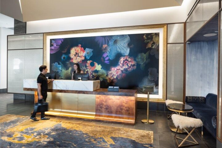 A man approaches a hotel reception desk with a floral mural behind it, while a receptionist stands ready to assist.