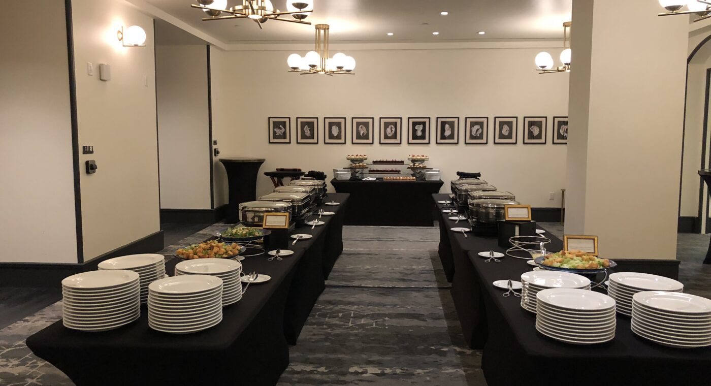 Buffet setup in a banquet room with neatly arranged plates, salads, and covered dishes on black tables under modern chandeliers.