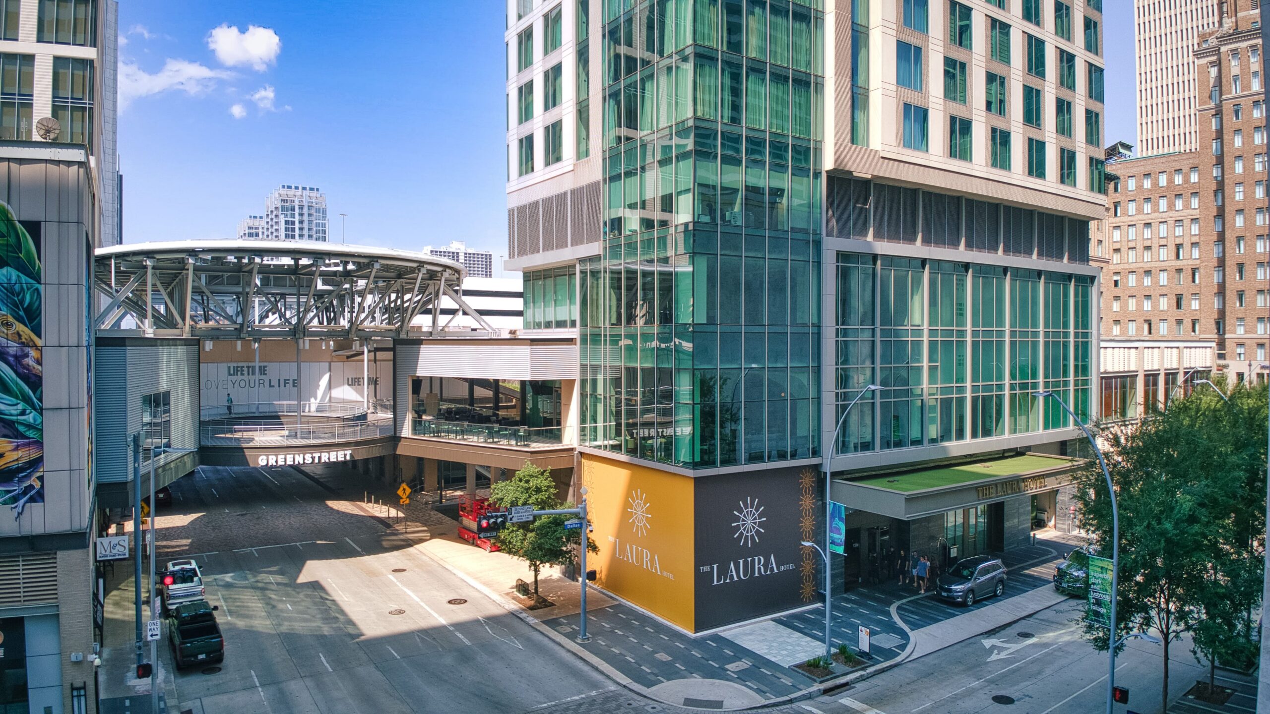 Modern city street with tall glass buildings, a skywalk, and businesses at street level, with minimal traffic on a sunny day.